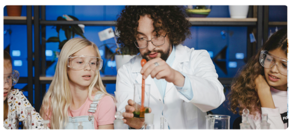 A teacher in a white lab coat fills a beaker as students in safety goggles look on